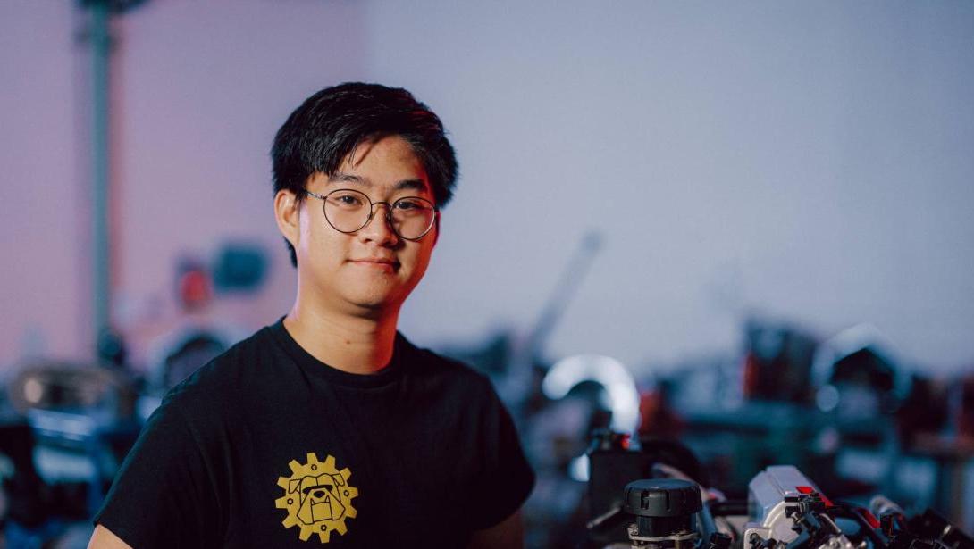 A Kettering student wearing a black shirt stands in the Car Lab alongside an engine component